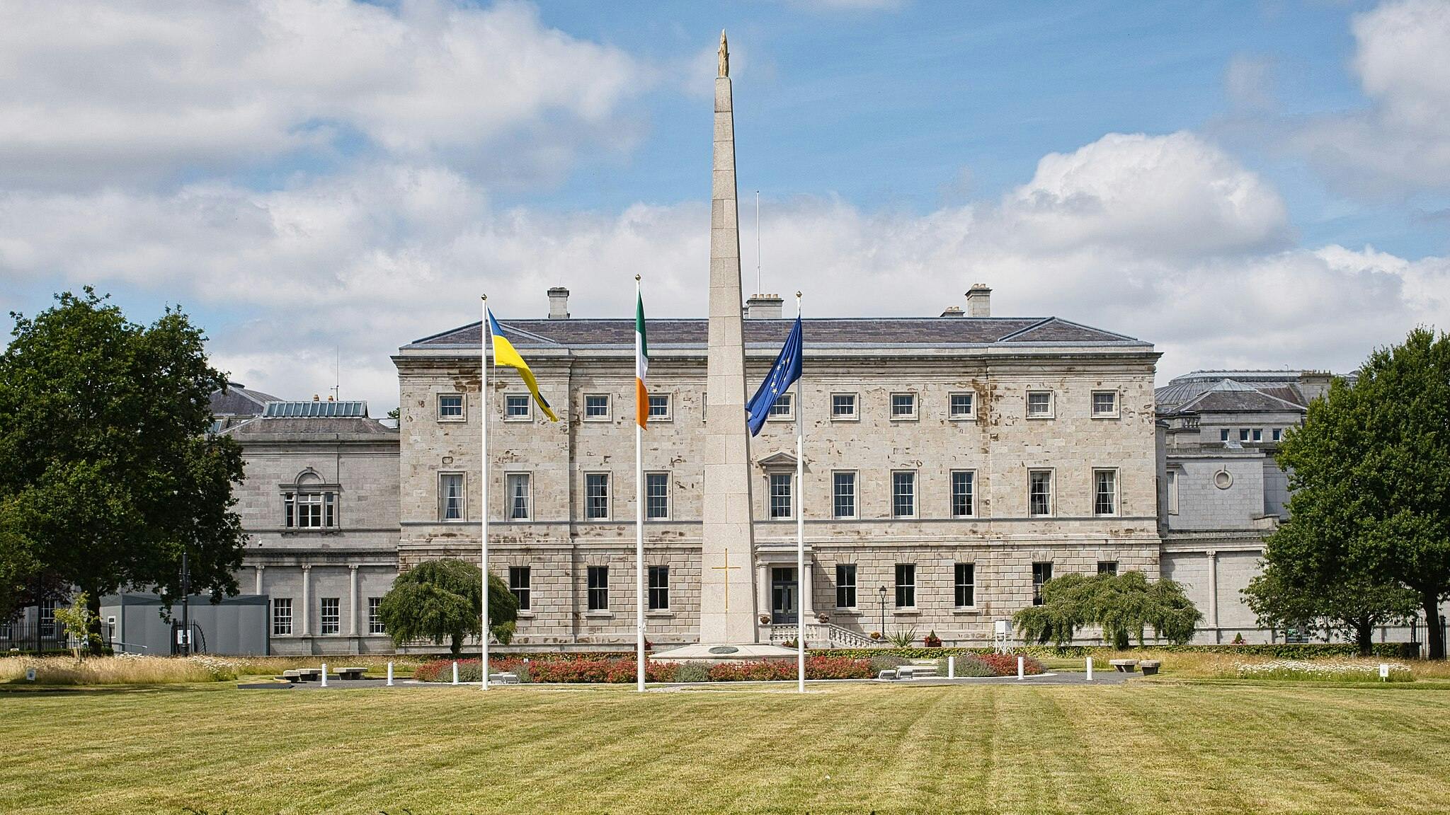 Historic stone building with flags and an obelisk in front, surrounded by a well-maintained lawn and a blue sky backdrop.