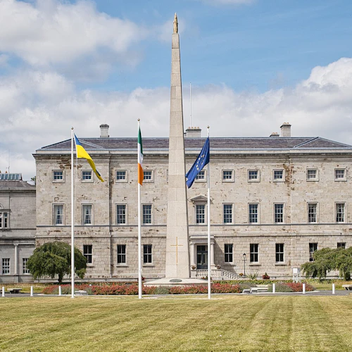 Historisk stenbyggnad med flaggor och en obelisk framför, omgiven av en välskött gräsmatta och en blå himmel.