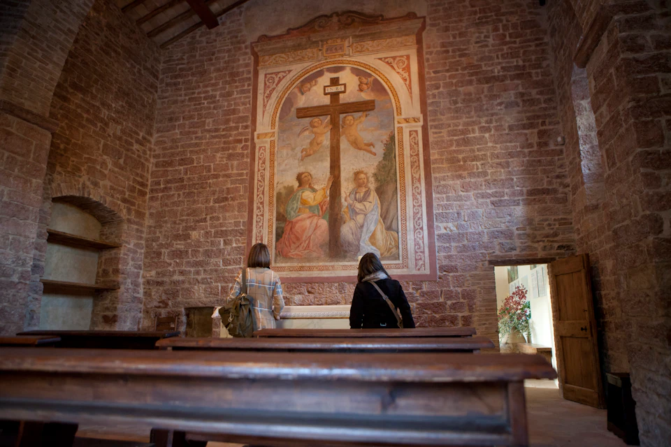 Two people in a church stand before a large mural of a cross flanked by two saints, with wooden pews and exposed brick walls.