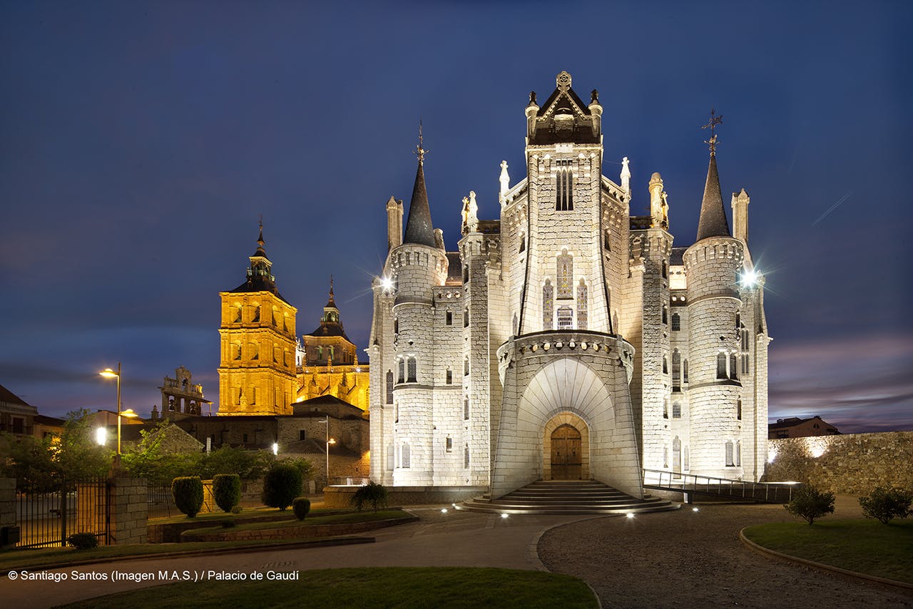Château de pierre illuminé avec de multiples tours, une entrée voûtée et un chemin menant à la porte sous un ciel crépusculaire.