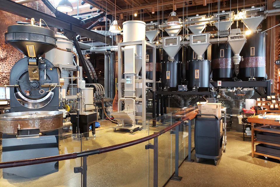 Industrial coffee roasting facility with large metal silos, pipes, and machinery, framed by glass barriers and exposed wooden ceiling.