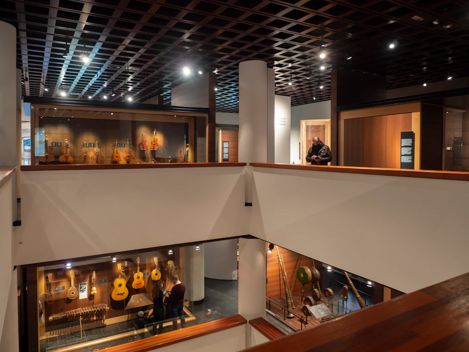 Interior of a multi-level museum with guitars and instruments displayed in cases. A few people are observing exhibits.