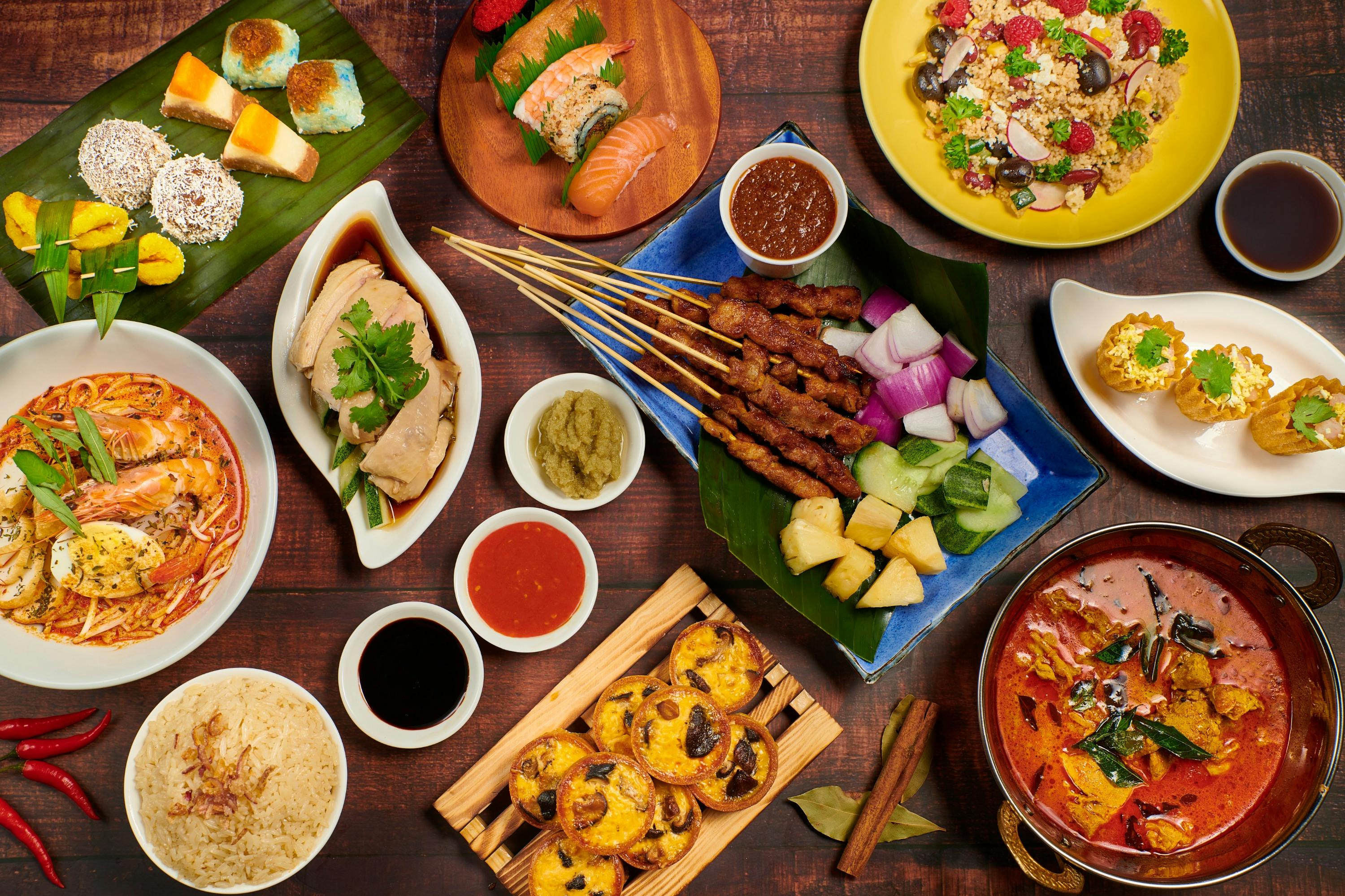 A variety of dishes on a wooden table including satay, rice, sushi, desserts, and curry, representing diverse cuisine.