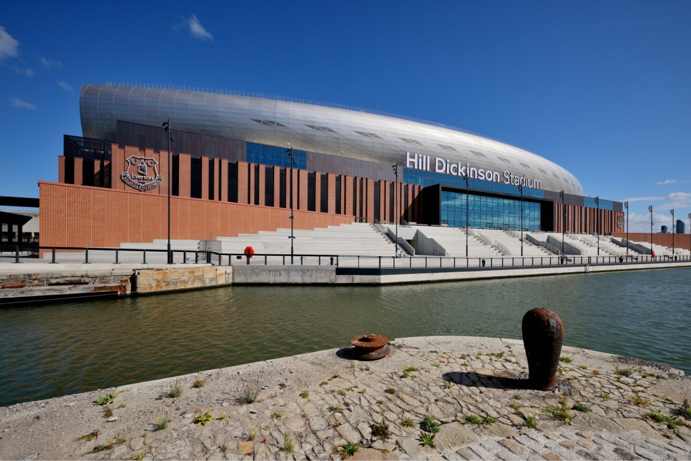 En stadionbyggnad märkt "Hill Dickinson Stadium" av en kanal, med klar himmel och stenbeläggning i förgrunden.