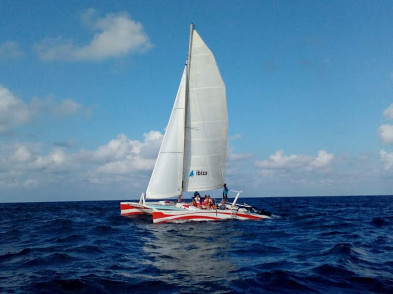 A catamaran with red and white hulls sails on the ocean under a blue sky with people sitting on the deck.