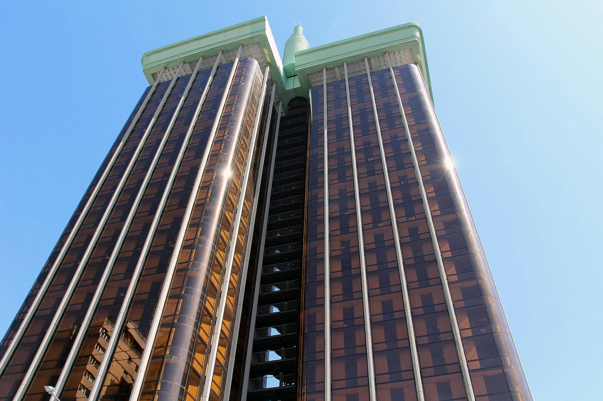 녹색 액센트가 있는 두 개의 높은 갈색 유리 마천루가 중간에 연결되어 맑고 푸른 하늘을 배경으로 낮은 각도에서 바라봅니다.