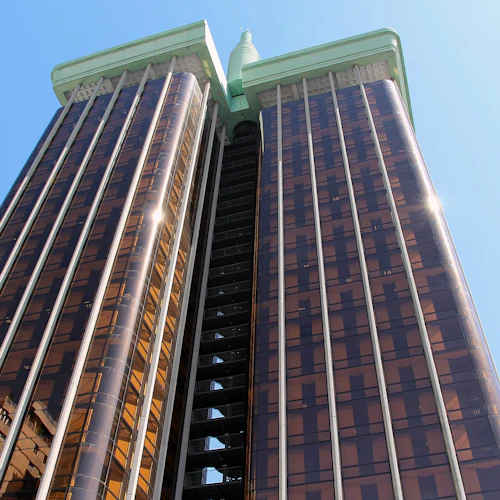 Två höga skyskrapor i brunt glas med gröna accenter, anslutna i mitten, sedda från en låg vinkel mot en klarblå himmel.
