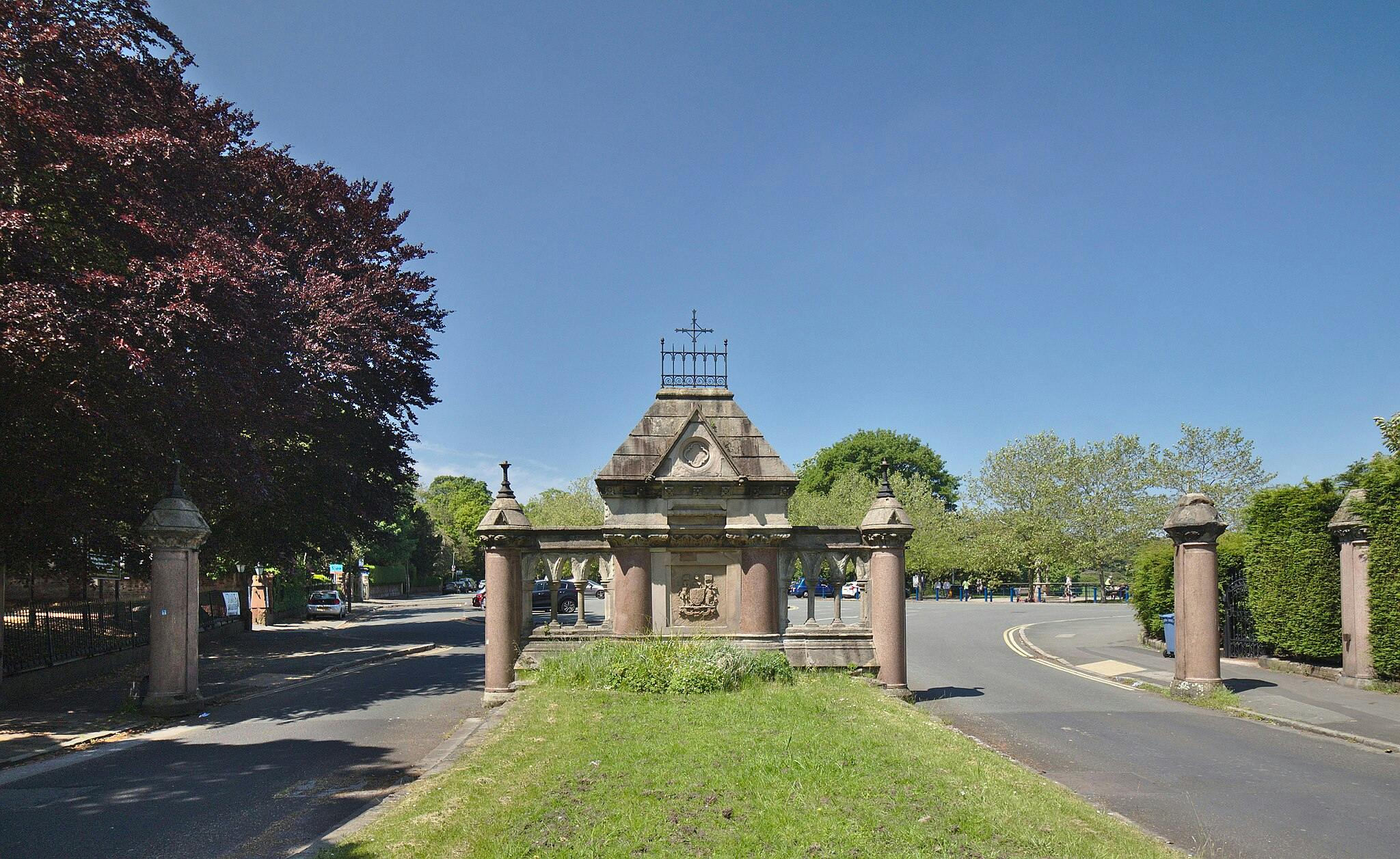 Περίτεχνη πέτρινη πύλη με κολώνες και διακοσμητικά γλυπτά, πλαισιωμένη από δέντρα σε μια ηλιόλουστη μέρα, που οδηγεί σε δρόμο.