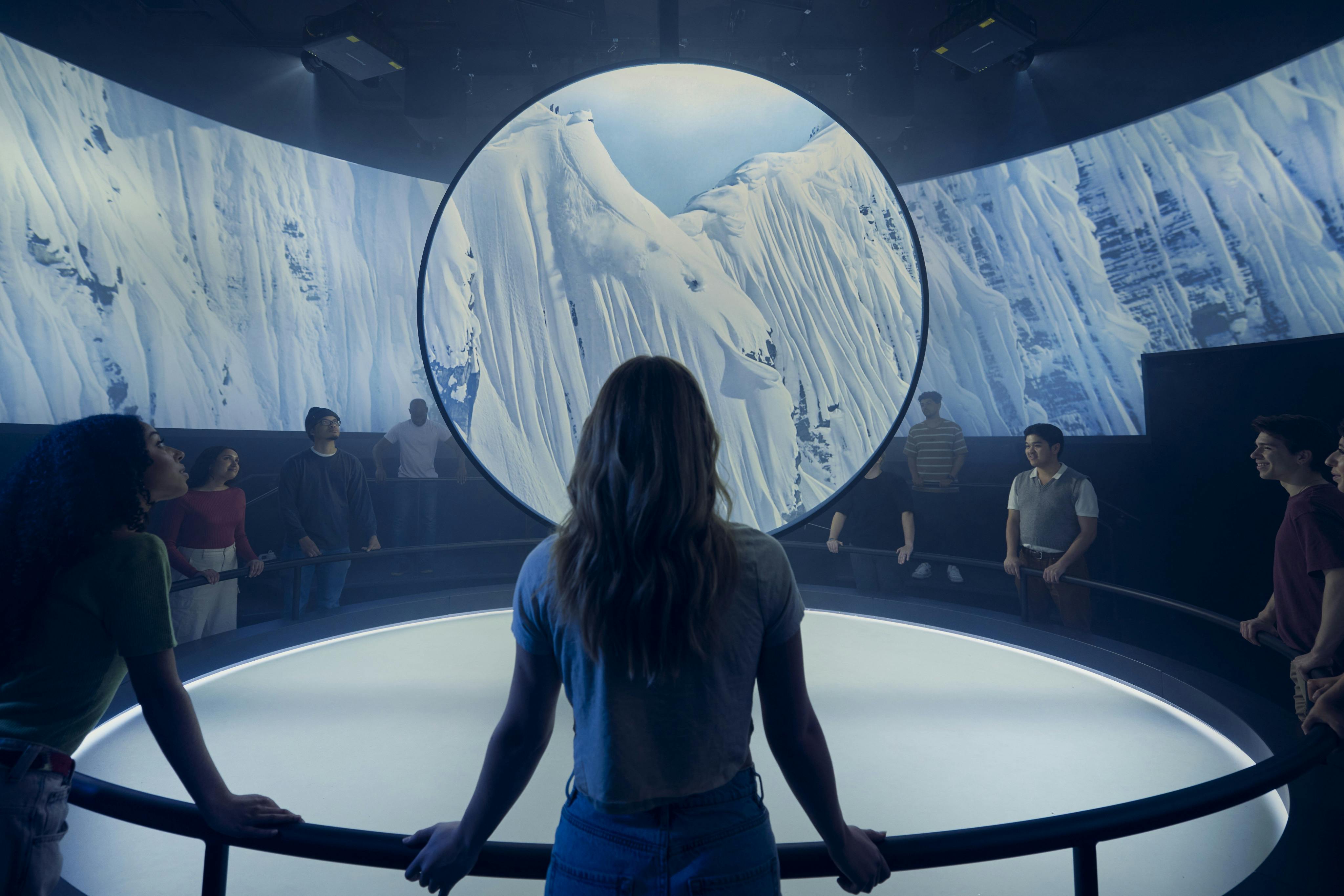 Een persoon staat voor een cirkelvormig scherm met besneeuwde bergen, omringd door anderen in een schemerige kamer.