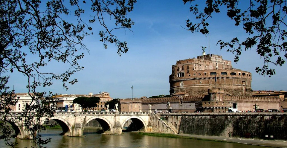 A historic circular fortress with a statue on top stands next to an arched stone bridge over a river, framed by tree branches.