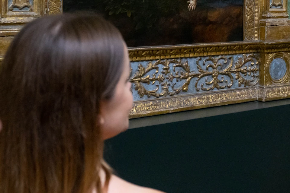 People viewing a framed painting in a museum, depicting a religious scene with several figures in a natural setting.