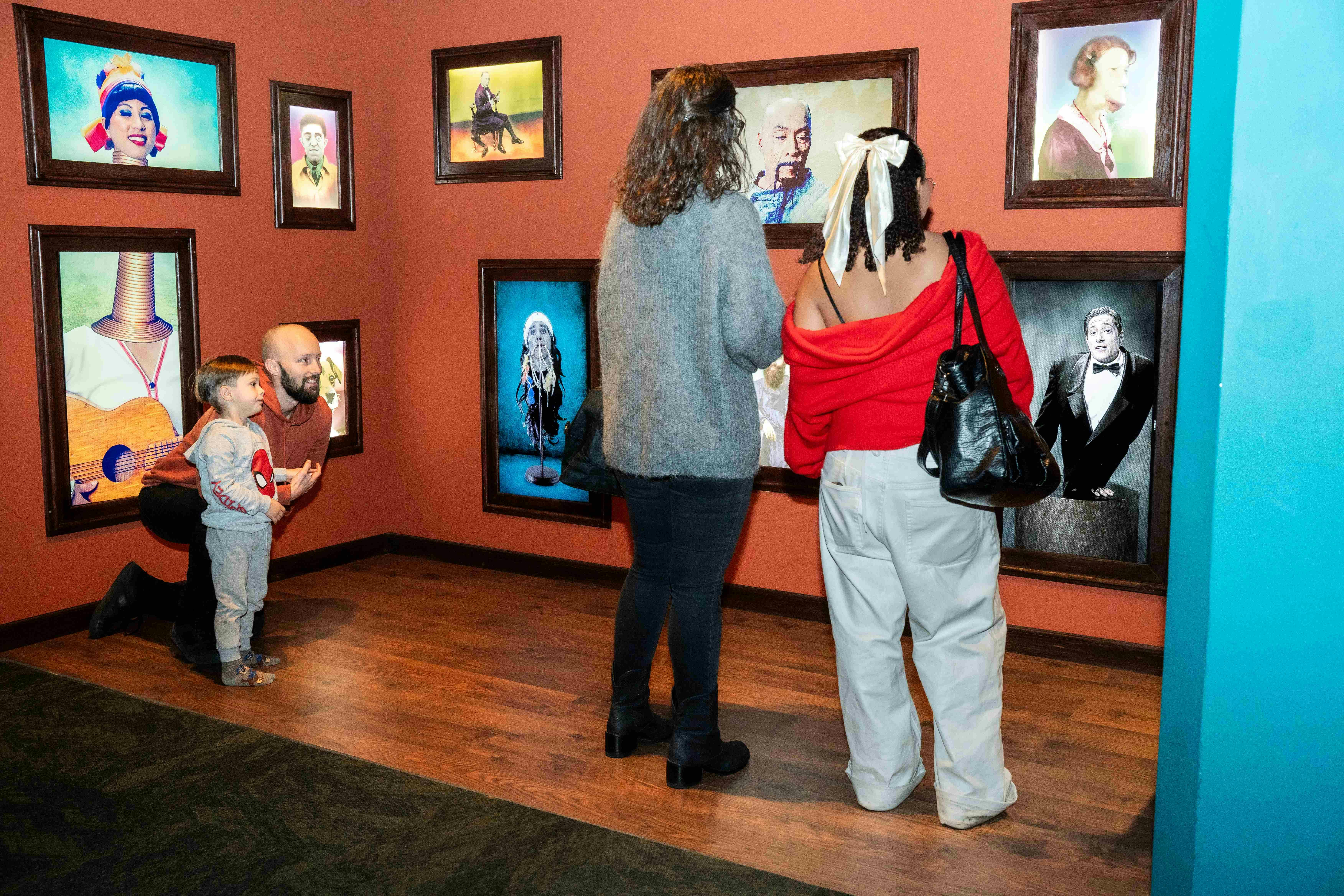 人们在画廊的橙色墙壁上欣赏带框的艺术品。 一个男人跪在一个孩子旁边，两个女人站在那里观察。