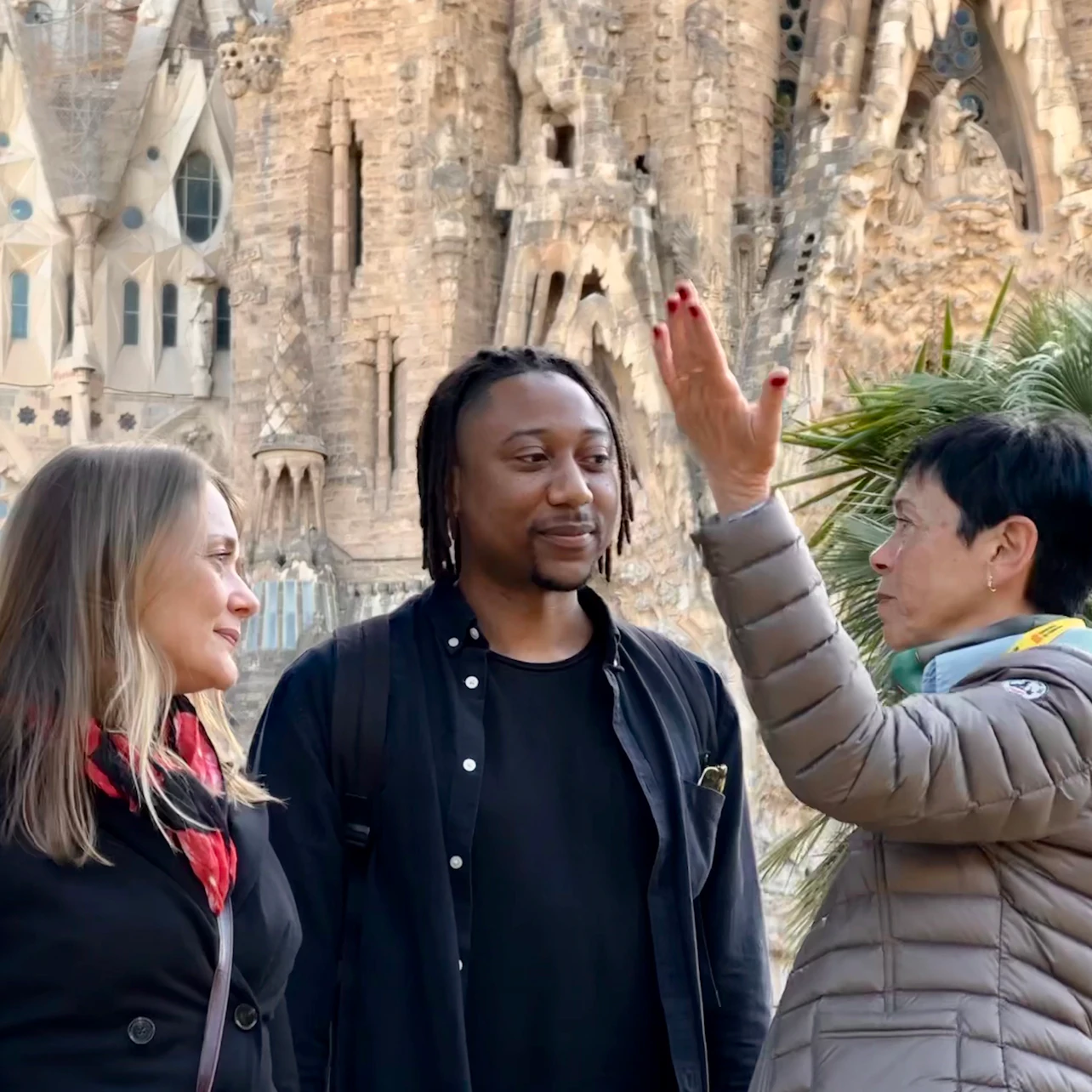 Three people are engaged in conversation with a detailed stone building in the background. One person is gesturing with their hand.