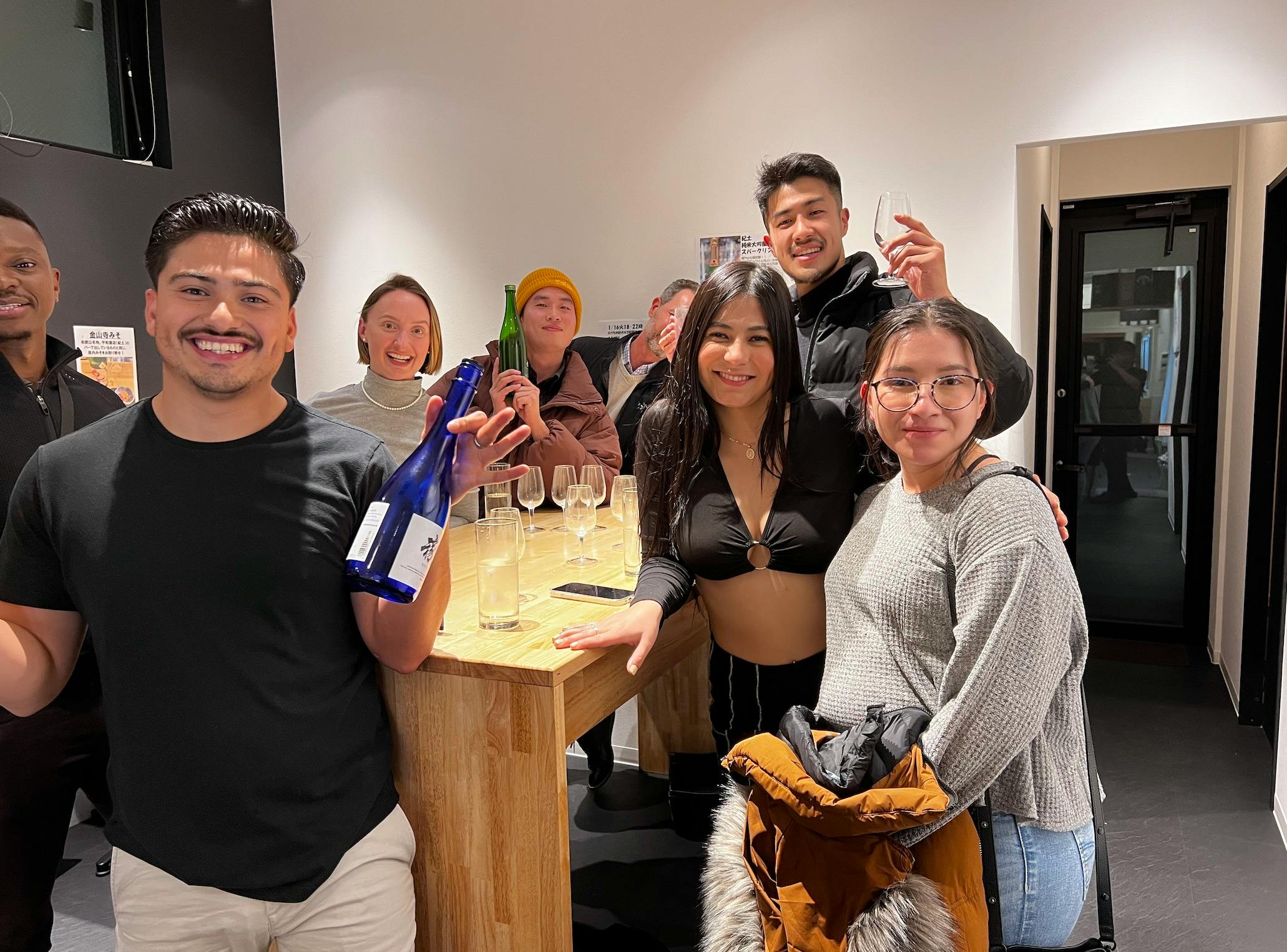 A group of people smiling and posing around a wooden table with bottles and glasses in a casual indoor setting.