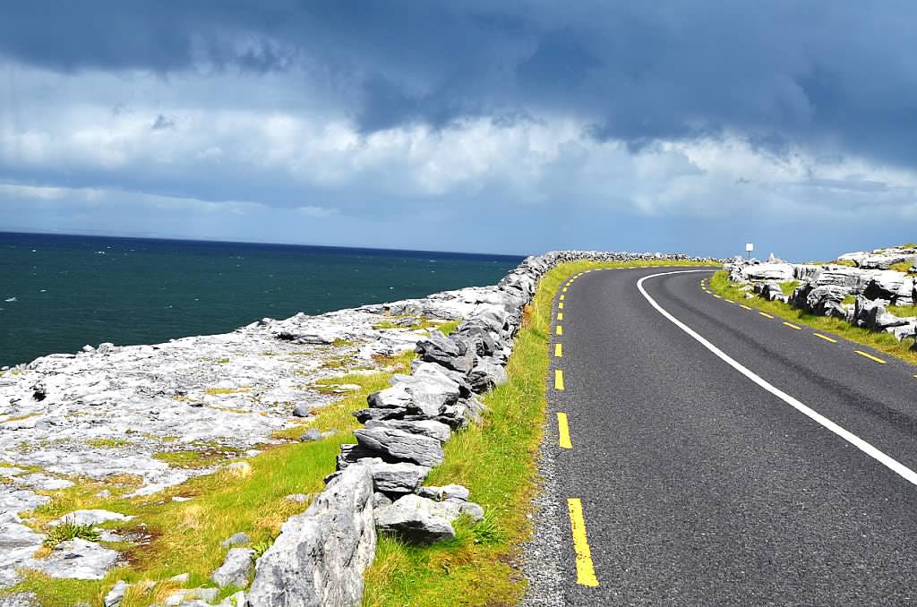 A coastal road with yellow dashed lines curves along rocky terrain beneath a cloudy sky, adjacent to a blue sea.
