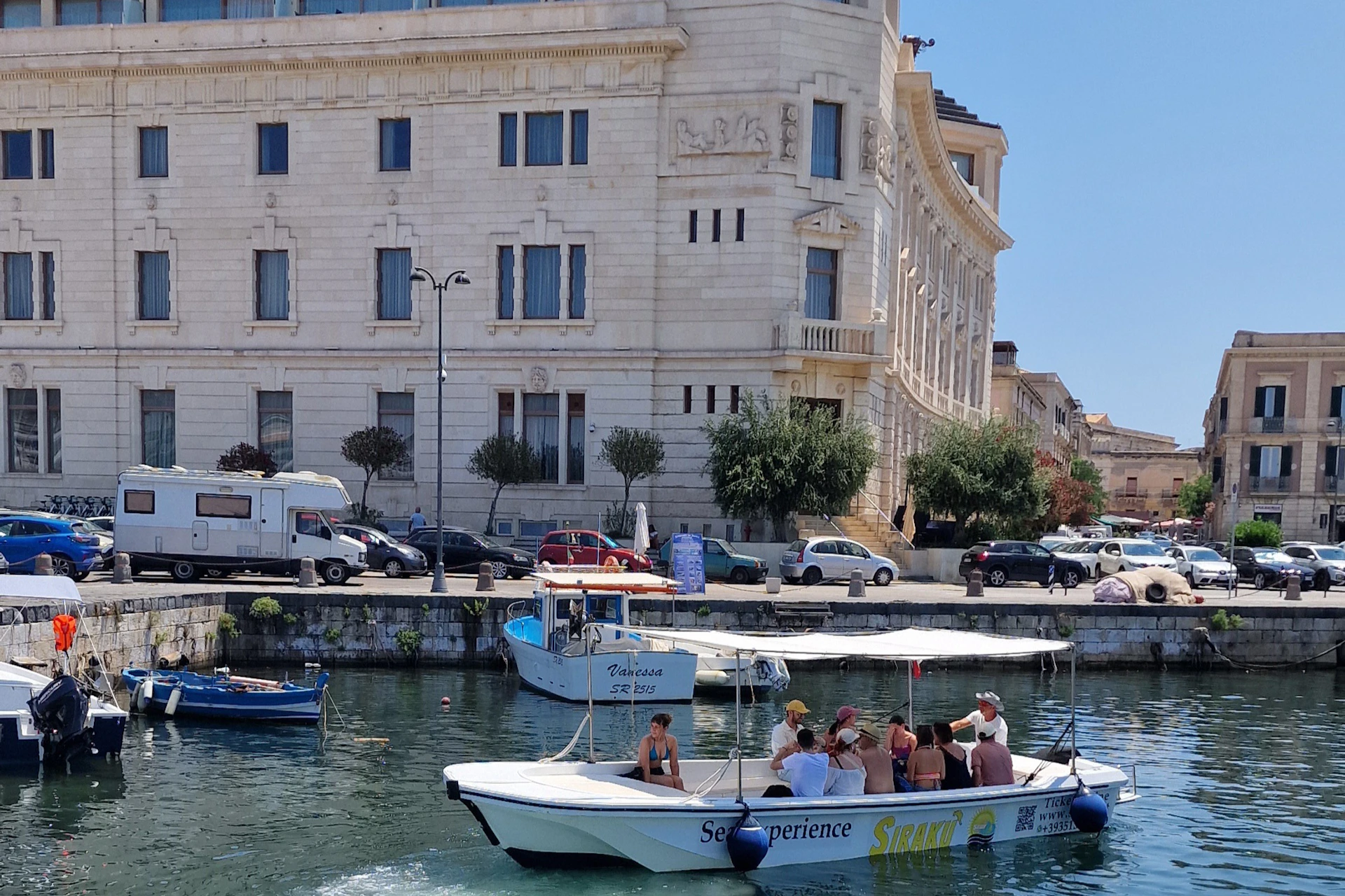 People on a tour boat labeled "Sea Experience" on a canal, with a large, ornate building and parked cars in the background.