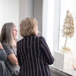 Två kvinnor står på ett museum och tittar på en detaljerad, utsmyckad guldskärm på en piedestal bakom glas.