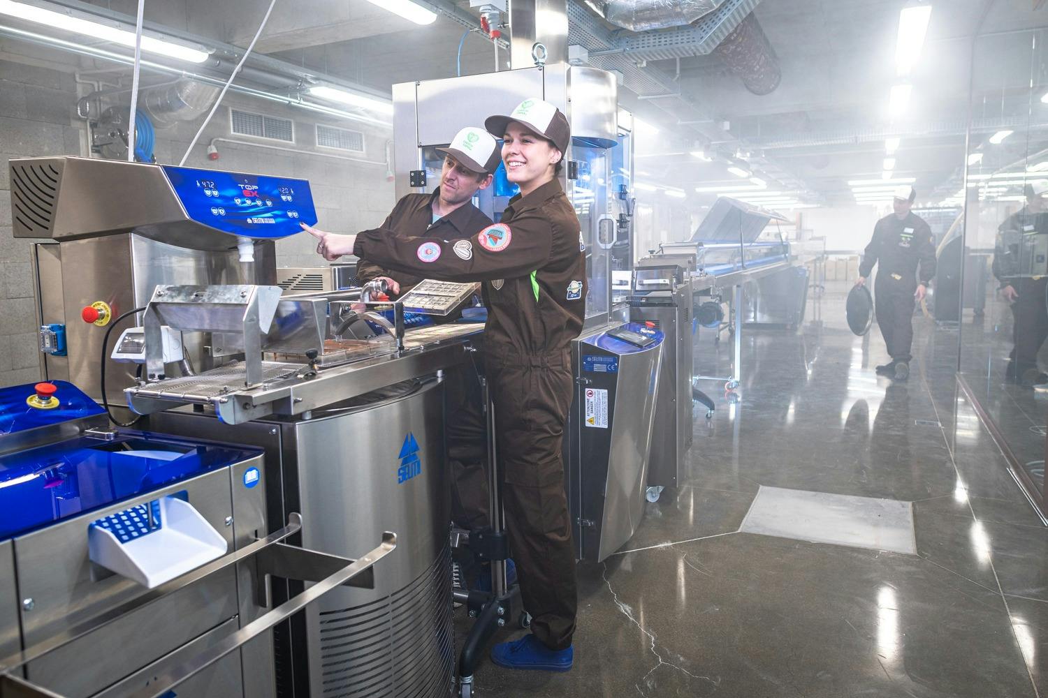 茶色のユニフォームとキャップを着た2人の労働者が工業施設で機械を操作し、別の労働者がバックグラウンドで歩いています。