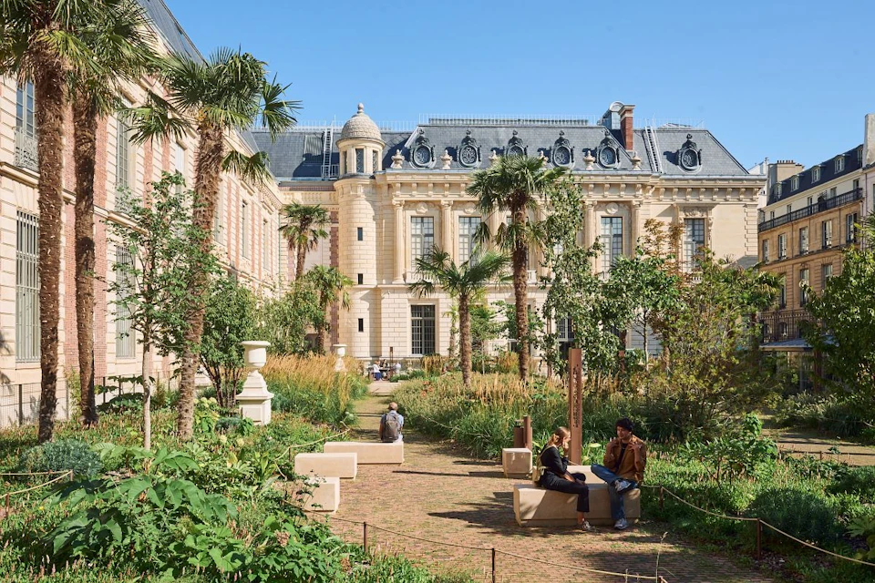 A garden with plants and palm trees in front of a classical building. People are sitting on benches and walking.