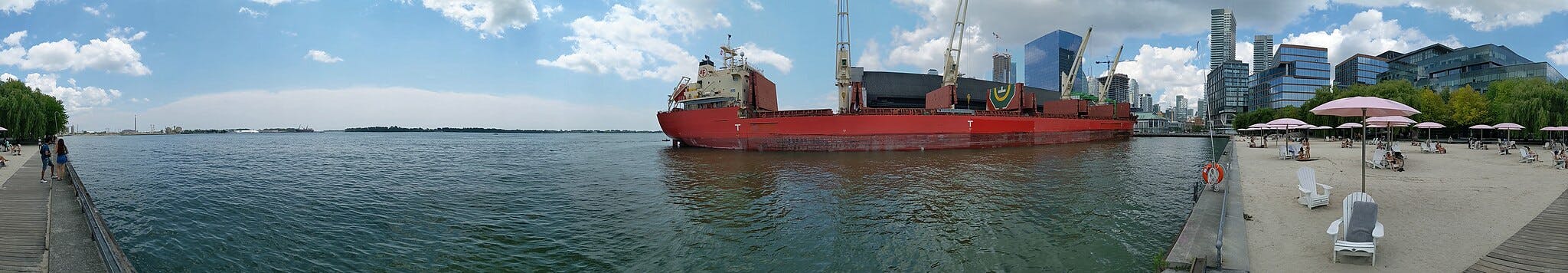 一艘红色货船停靠在现代城市建筑附近；人们在粉红色雨伞下的沙滩上放松身心。