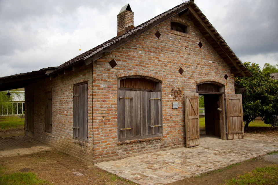 A brick building with wooden shutters and door, featuring a chimney and small loft window, under a cloudy sky.