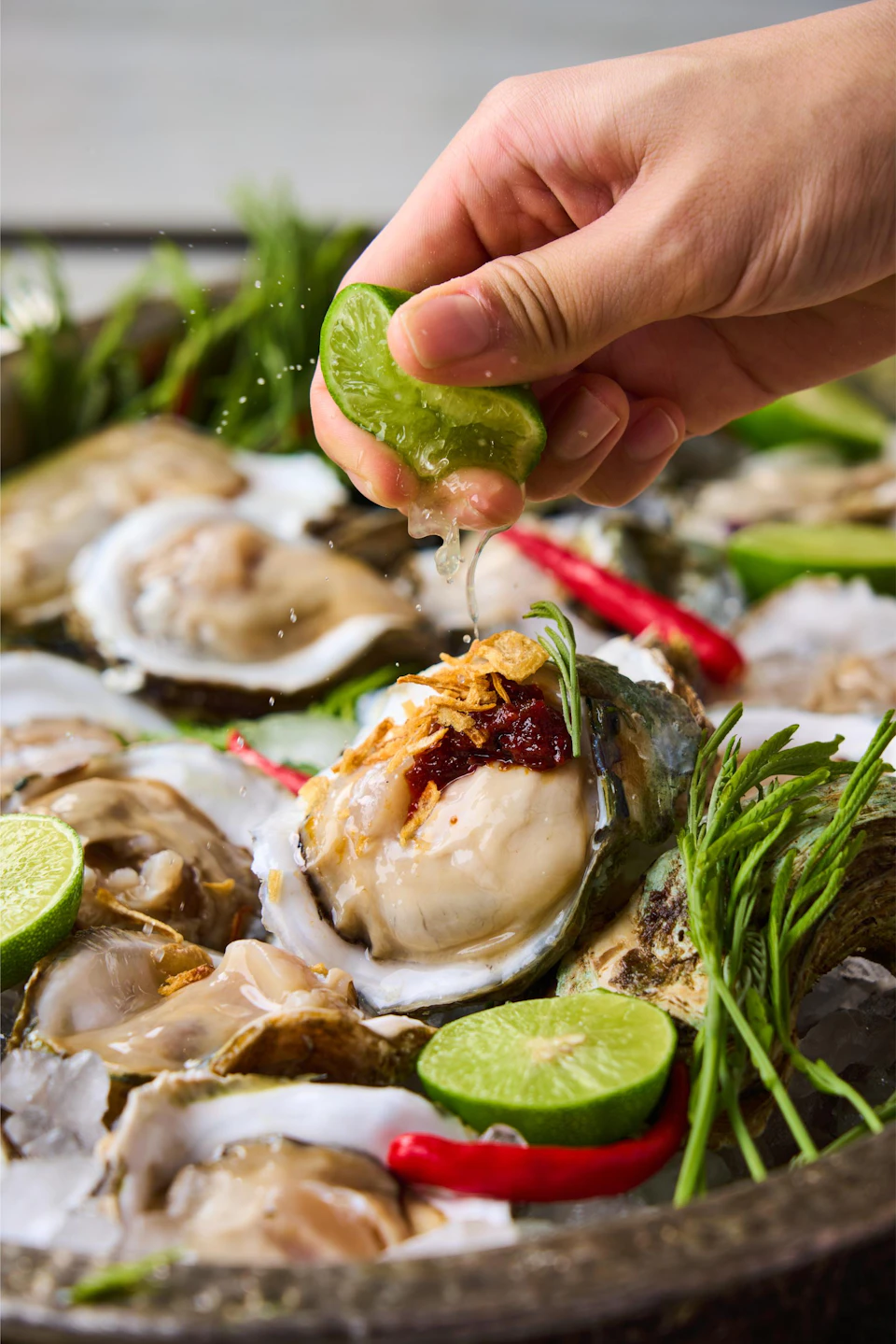 A hand squeezing lime juice over a plate of fresh oysters garnished with red sauce, fried onions, and herbs.