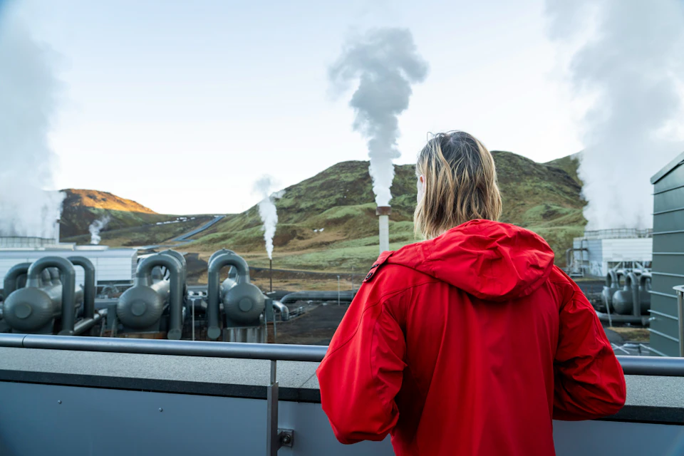 A person in a red jacket looks at a geothermal power plant with steam rising against a backdrop of grassy hills.