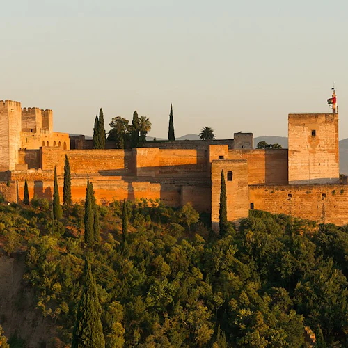 En historisk fästning med höga murar och torn, belägen på en kulle omgiven av täta gröna lövverk och badad i varmt solljus.