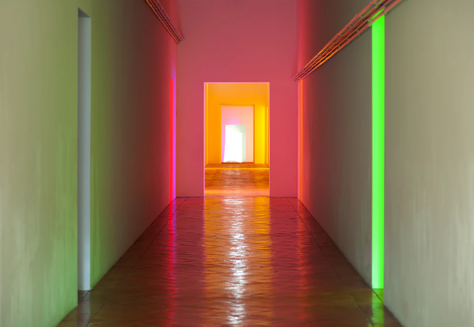 A hallway with colorful lighting in pink, green, and yellow reflecting on the walls and floor, leading to an open doorway.