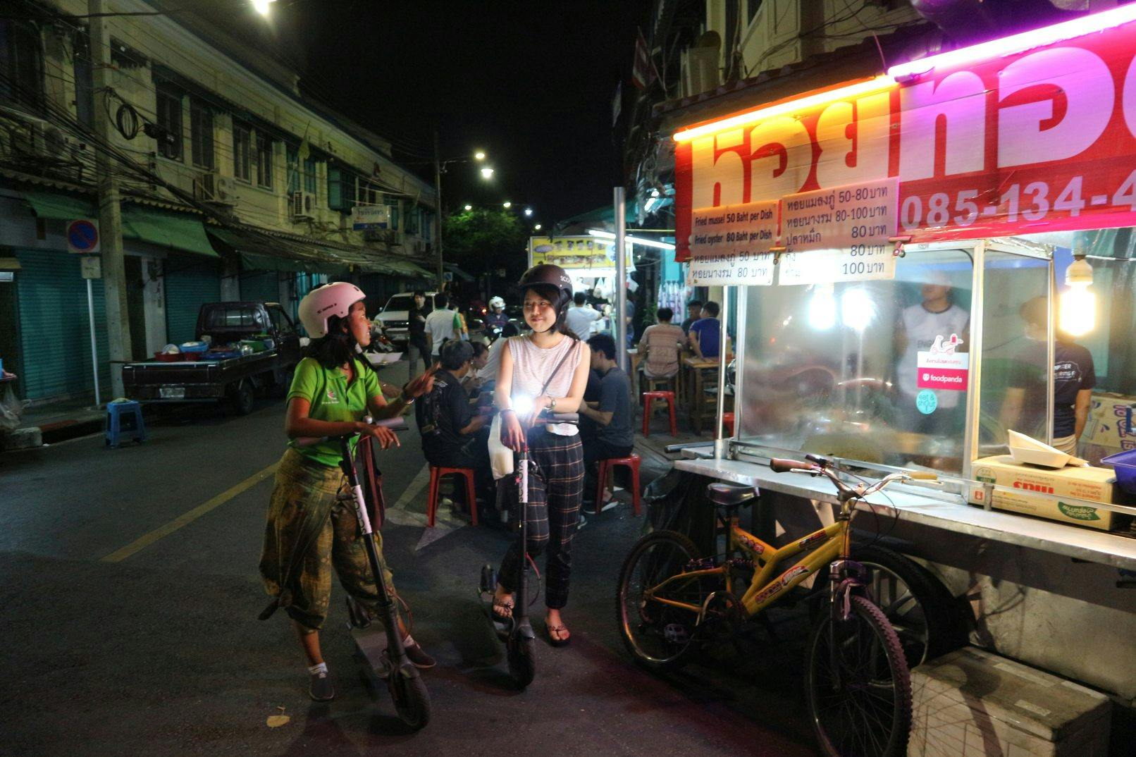 Two people wearing helmets stand with electric scooters on a nighttime street near a food stall with neon signage.