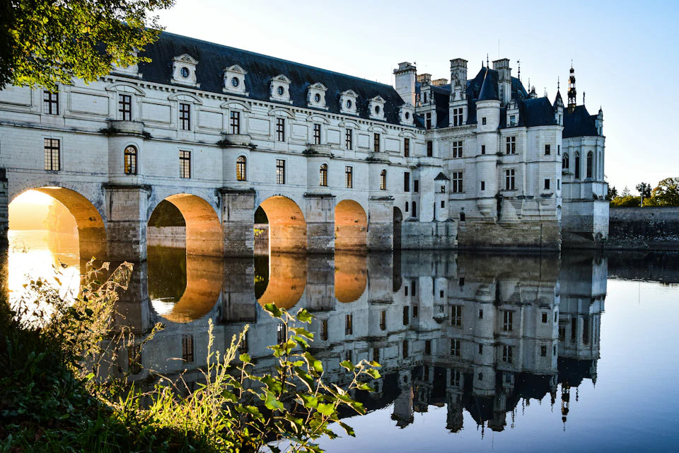 A historic building with arches spans over a reflective body of water, surrounded by greenery under a clear sky.