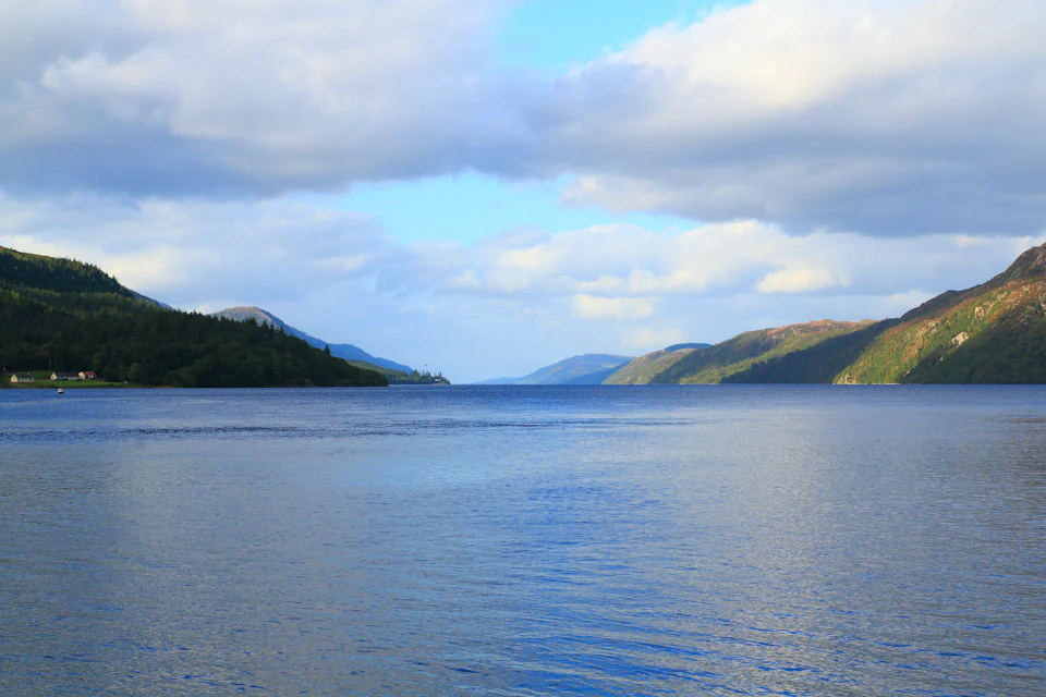 A calm lake surrounded by tree-covered hills under a partly cloudy sky.