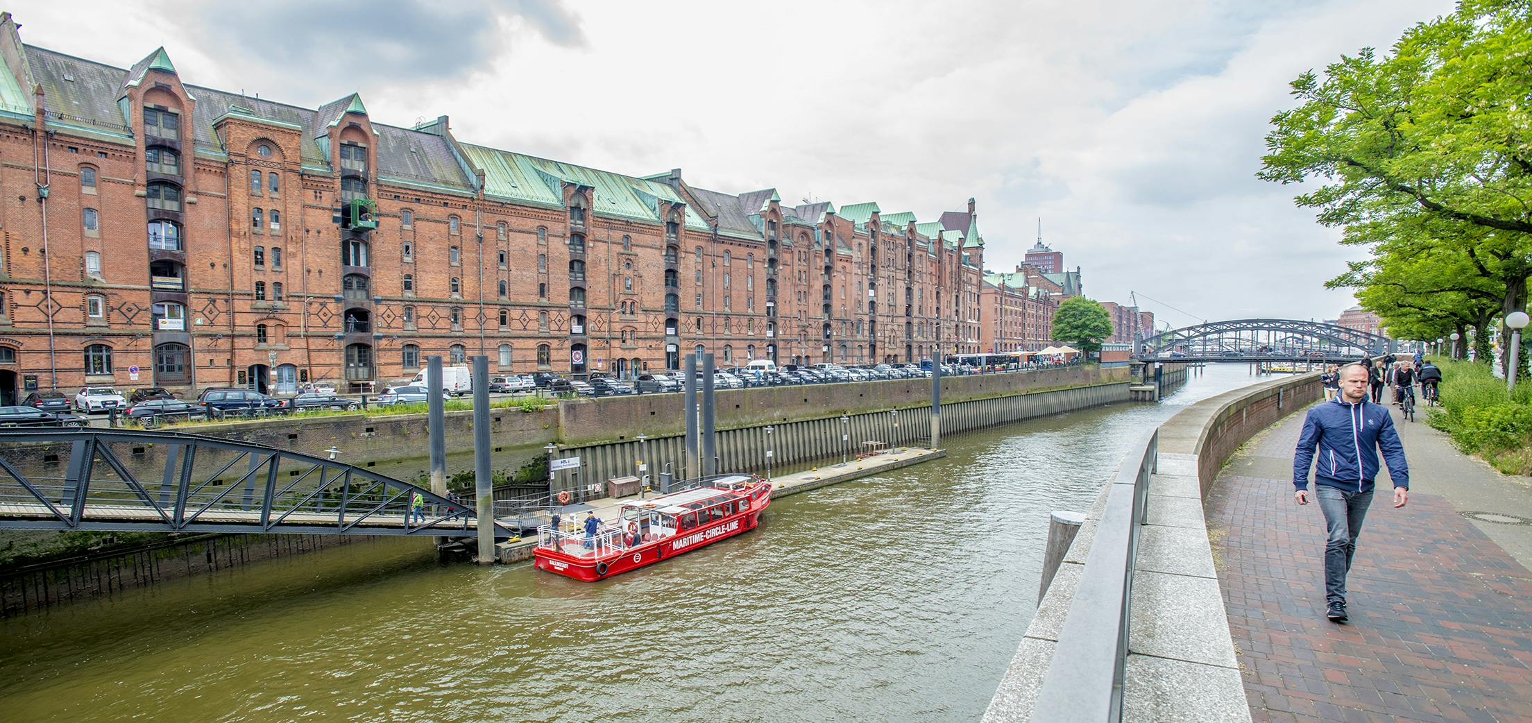 Un homme marche sur un sentier bordé d'arbres et de bâtiments historiques, avec des ponts au loin, sous un ciel nuageux.