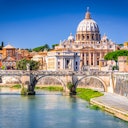 A view of St. Peter's Basilica in Vatican City with the Tiber River and a stone bridge in the foreground under a clear blue sky.