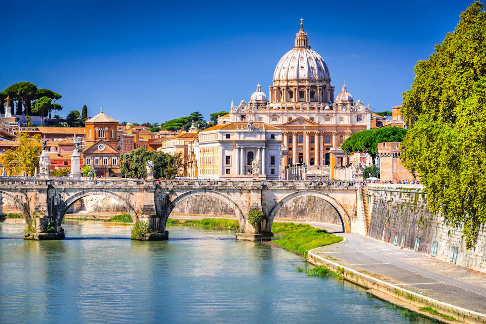 A view of St. Peter's Basilica in Vatican City with the Tiber River and a stone bridge in the foreground under a clear blue sky.