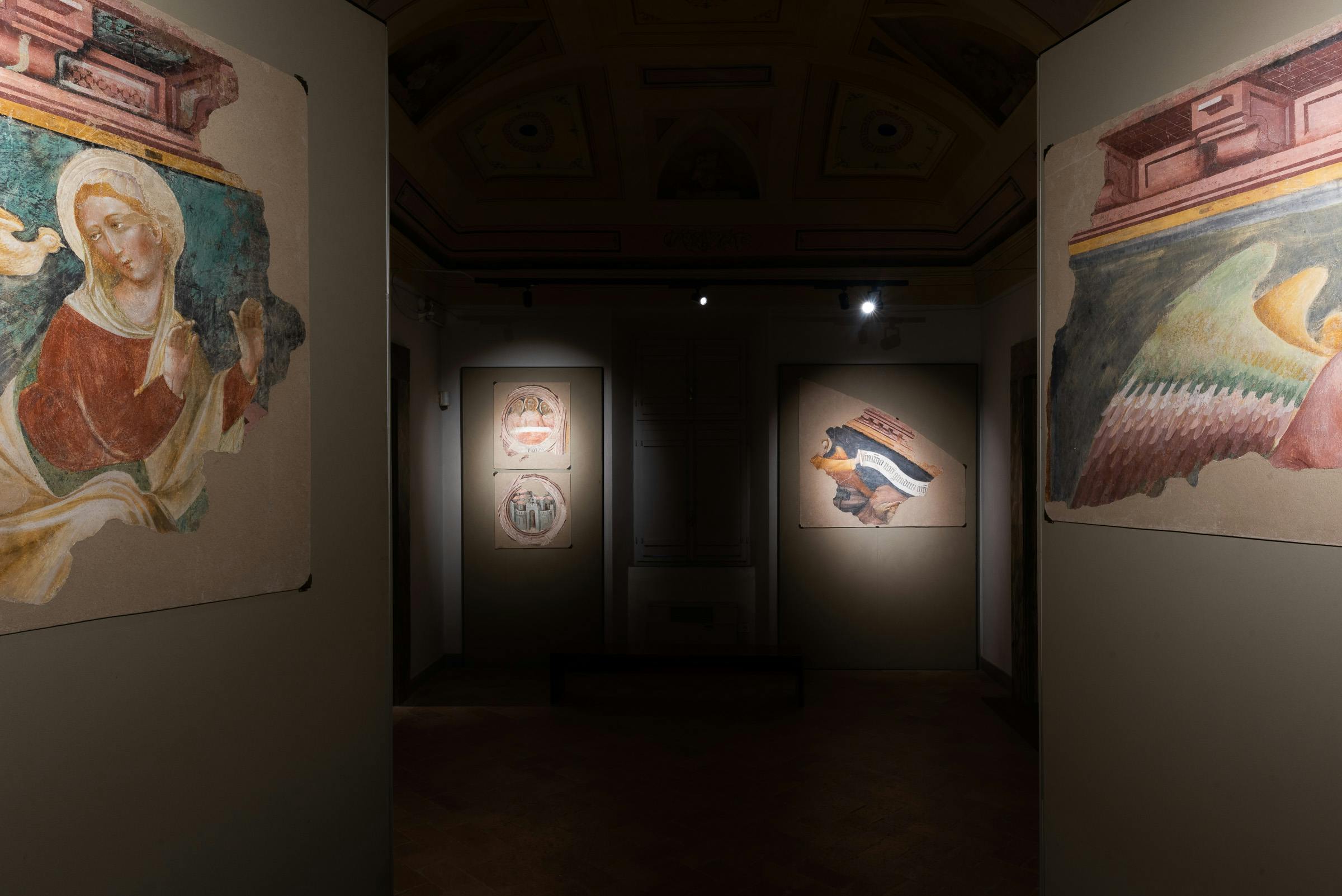 Galleria poco illuminata che espone antichi affreschi alle pareti, con faretti che evidenziano pezzi specifici su sfondi beige.