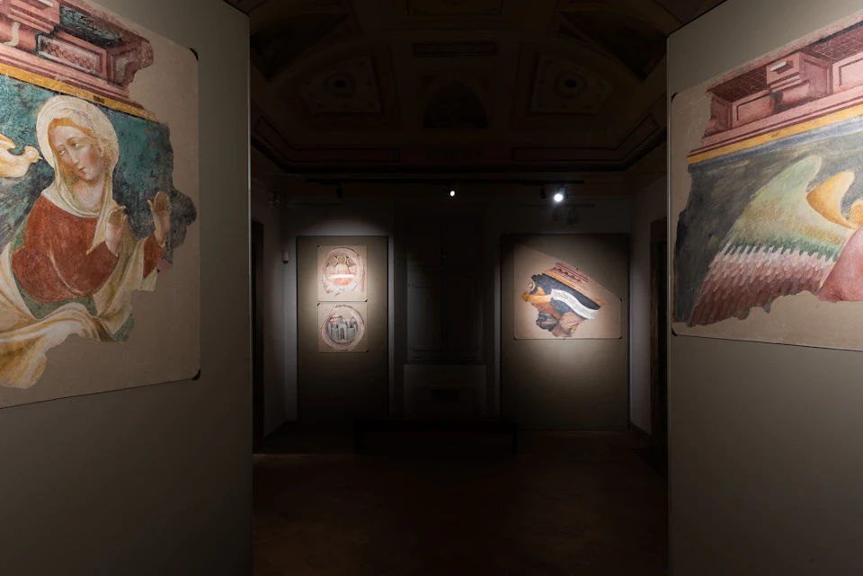 Dimly lit gallery displaying ancient frescoes on walls, with spotlights highlighting specific pieces on beige backgrounds.
