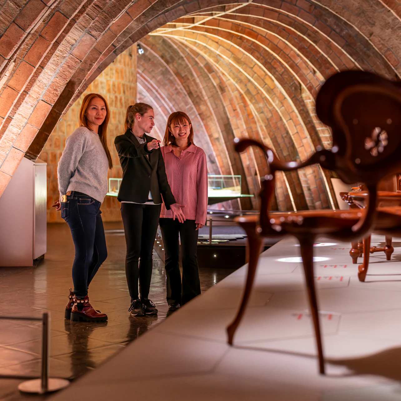 Three women admiring exhibits in a warmly lit, arched room with displayed furniture pieces.
