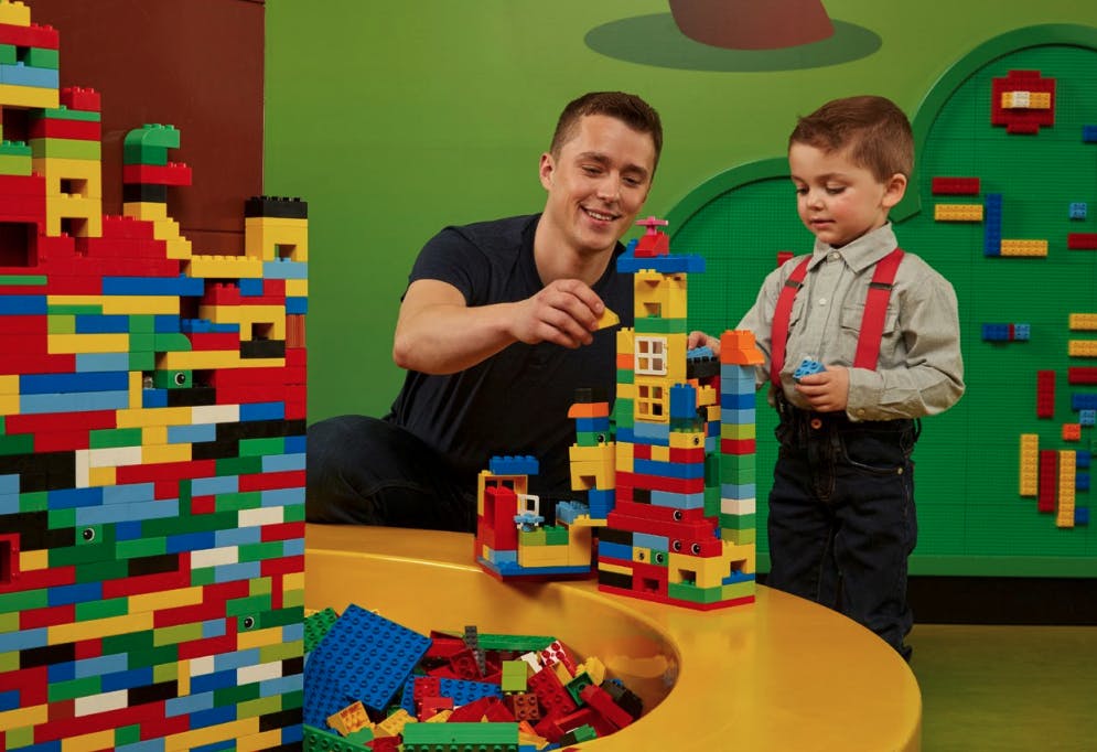 男性と男の子は、緑色の背景の黄色いテーブルの上にレゴのレンガでカラフルな構造を構築します。