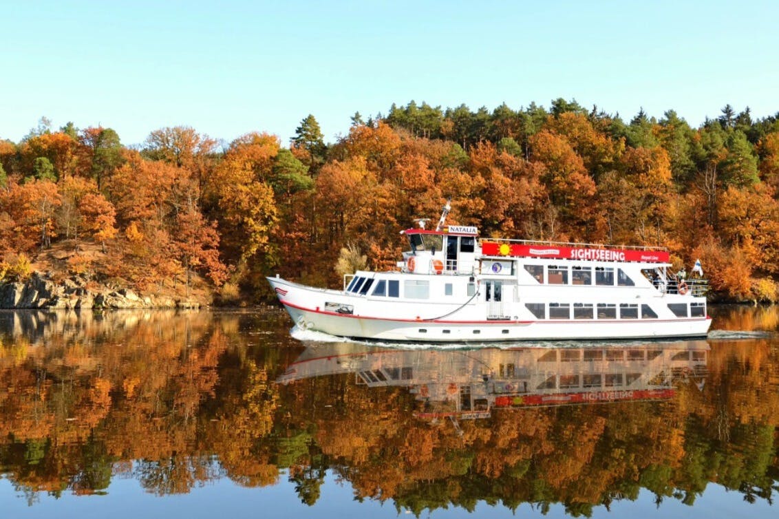 Biała łódź krajoznawcza płynie po spokojnej wodzie, odbijając jesienne drzewa z pomarańczowymi i żółtymi liśćmi w tle.