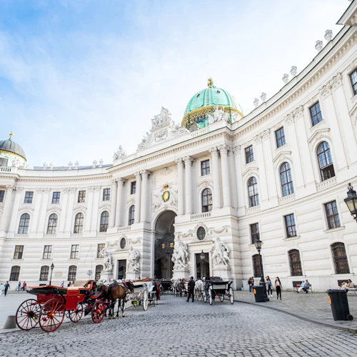 Halvcirkelformad vit byggnad med gröna kupoler, statyer, hästdragna vagnar och människor som går på en kullerstensgata.