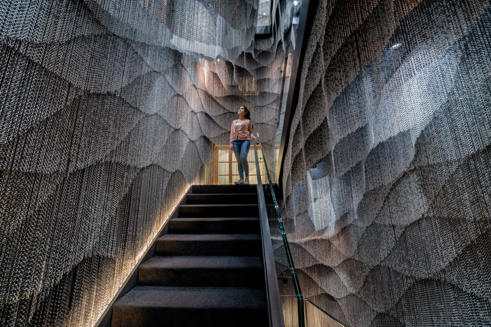 A woman stands on a staircase surrounded by textured, wave-like walls made of chain-like materials in a modern interior.