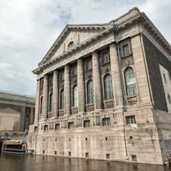 Två stora neoklassiska byggnader med pelare, bredvid en kanal, under en molnig himmel. Konstruktionsmaterial synliga vid vattnet.