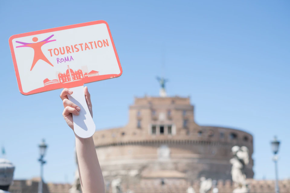 A hand holding a sign reading 'Touristation Roma' in front of Castel Sant'Angelo against a blue sky.