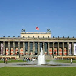 En neoklassisk byggnad med höga pelare, stora banderoller, en fontän framför och människor som går på en gräsbevuxen plaza under en klarblå himmel.