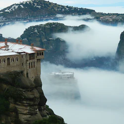 Ett kloster sitter på en klippa bland dimmiga moln med bergig terräng i bakgrunden.