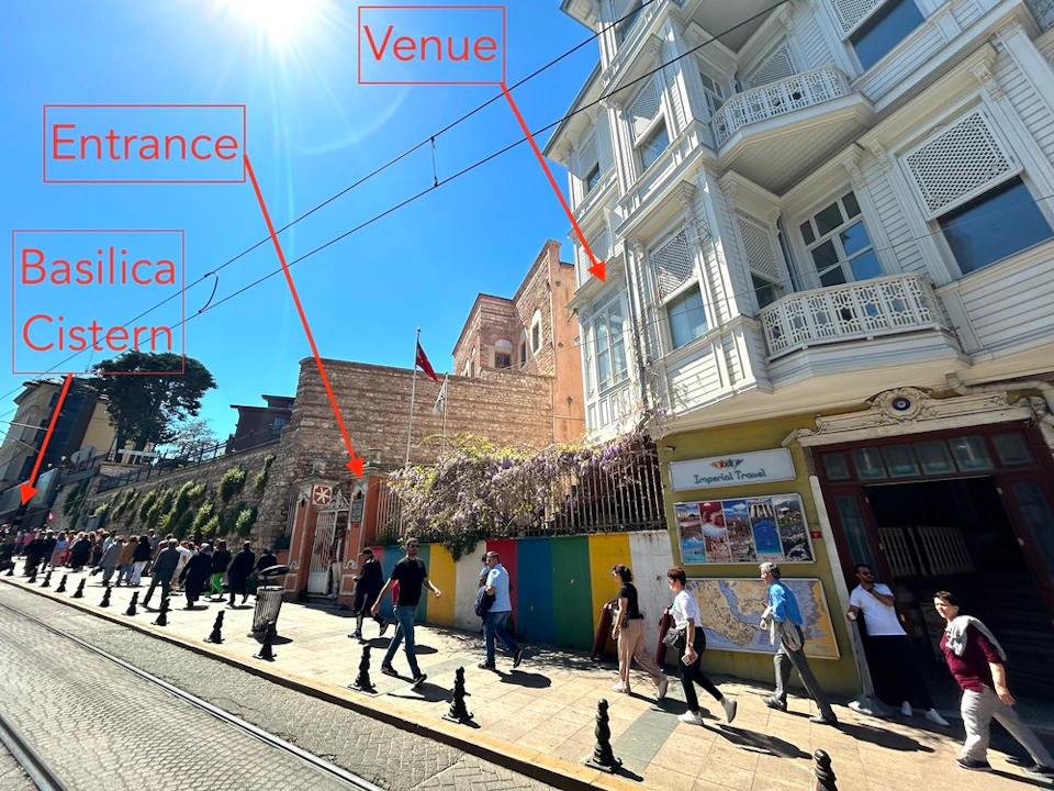 People walking along a cobblestone street on a sunny day, with a historical building and a sign reading "Venue" pointing upwards.
