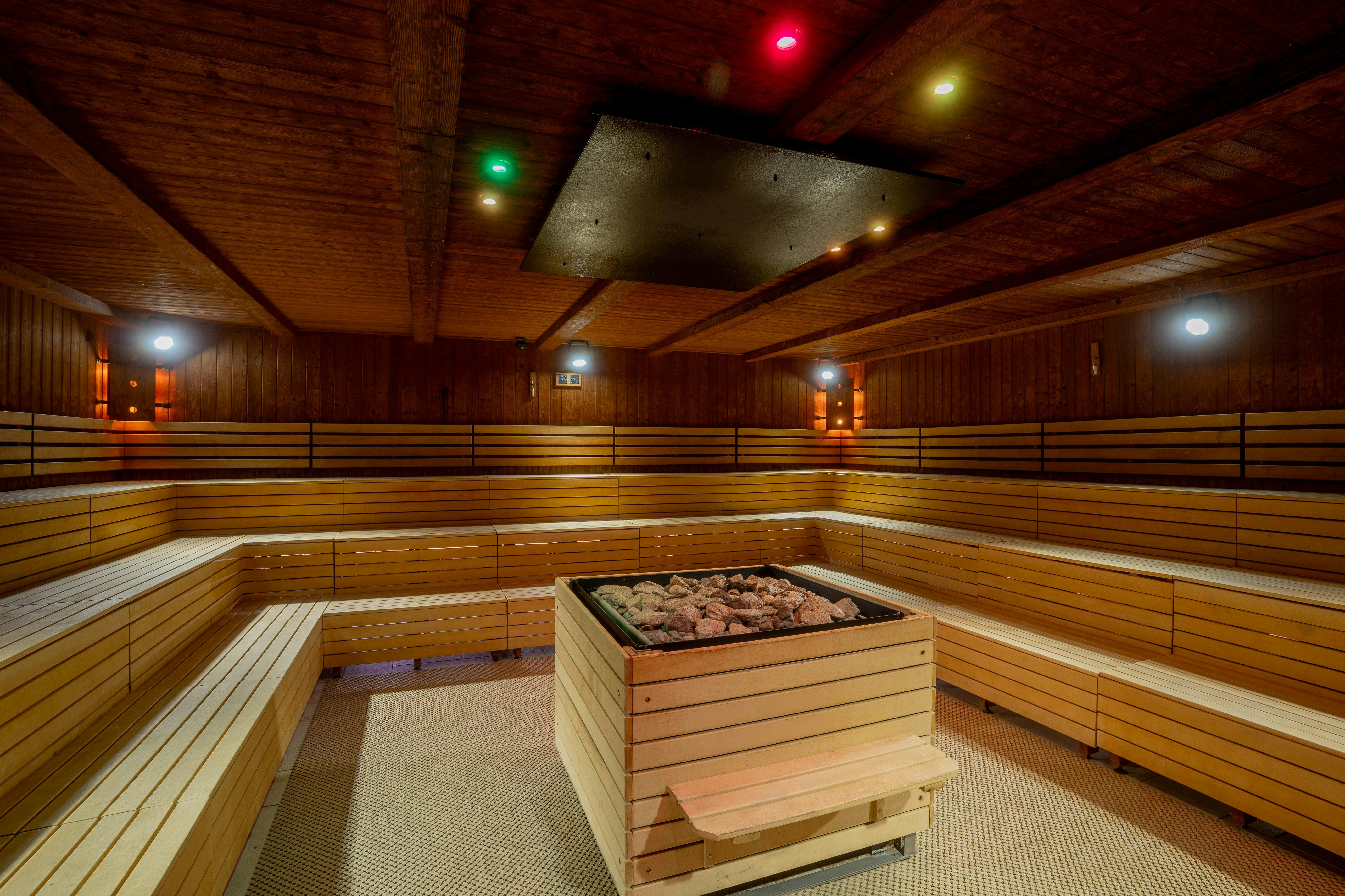 A wooden sauna room with bench seating along the walls and a central heater filled with stones. Colorful ceiling lights are visible.
