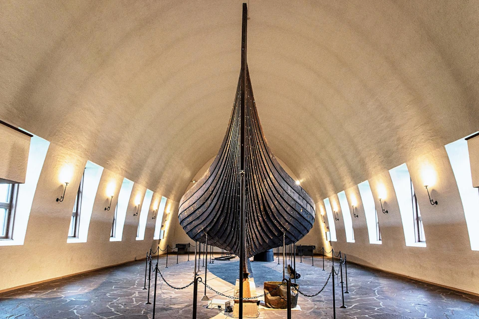 A large, historic Viking ship displayed in a museum with a high arched ceiling and several windows.