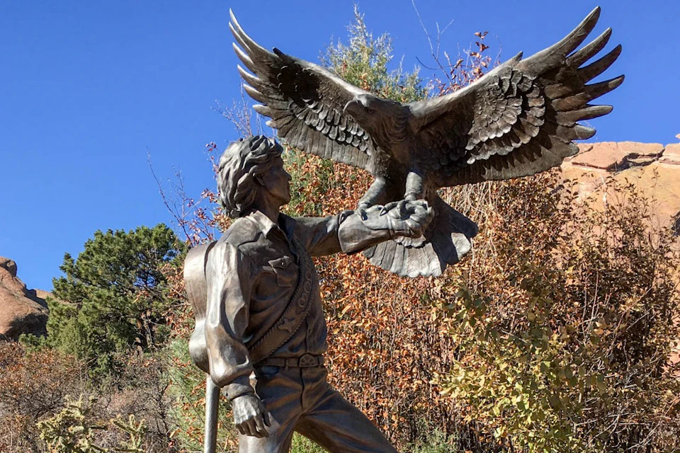 Bronze statue of a person holding a perched eagle, with trees and a clear blue sky in the background.